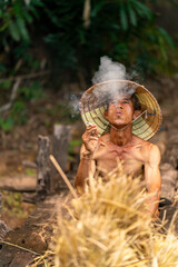 Photo of an asian old male farmer village man wearing conical hat taking a break while delivering some dry grasses for animal feeds rest sitting on a wooden bridge smoke a local tobacco