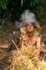 Photo of an asian old male farmer village man wearing conical hat taking a break while delivering some dry grasses for animal feeds rest sitting on a wooden bridge smoke a local tobacco