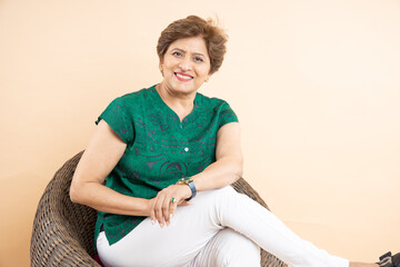 Portrait of smiling senior Indian woman sitting relaxing on chair isolated on beige background....