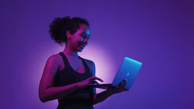 Online fitness. Happy woman. Digital technology. Positive lady in sportswear typing laptop holding hands posing neon light purple white background.