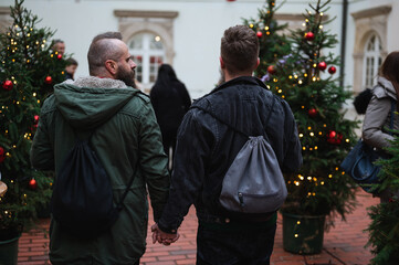Fashionable male gay couple walking away and holding hands  on Christmas advent outdoor. Hipster homosexuals together in public, stylish in winter jackets and rucksacks.