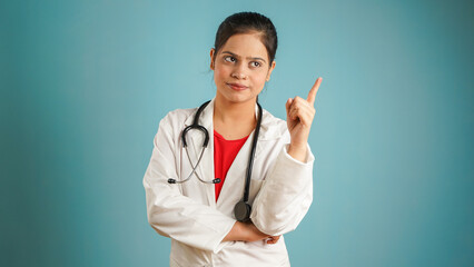 Portrait of a young female doctor pointing finger at copy space, Cheerful Asian Indian woman doctor in apron and stethoscope showing empty copy space isolated over blue studio background