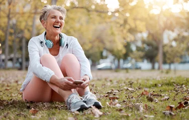 Poster Health, nature and senior woman in park with phone and headphones to relax during fitness workout. Music, happiness and old lady laughing, sitting on ground in garden after running exercise in autumn © Anela/peopleimages.com