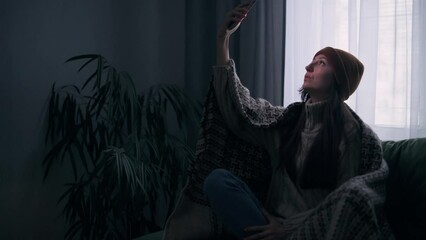 A young woman is trying to catch a connection during a power outage. The female sits on a couch in a dark room with no electricity, no communication, and no heating.