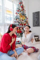 Two young asian female lady friends in a room listening to new songs on their headphone via their digital tablet celebrate Christmas together