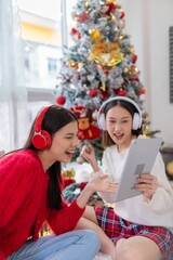 Two young asian female lady friends in a room listening to new songs on their headphone via their digital tablet celebrate Christmas together