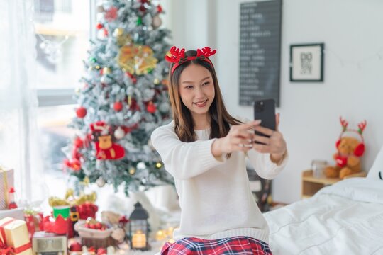 Cute beautiful young asian lady woman wearing christmas reindeer headband posing cheerfully taking selfie pictures with her mobile phone beside a big Christmas tree with lots of decoration ornaments