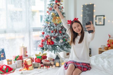 Cute beautiful young asian lady woman wearing reindeer headband selfie and video call with mobile phone posing in front a big Christmas tree with lots of decoration lights gift box and ornaments