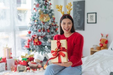 Obraz na płótnie Canvas Cute beautiful young asian lady woman wearing reindeer headband holding a gold color gift box with red ribbon posing in front a big Christmas tree with lots of decoration lights gift box and ornaments