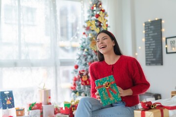 Cute beautiful young asian lady woman wearing reindeer headband holding a green color gift box with bells and posing in front a big Christmas tree with lots of decoration lights gift box and ornaments