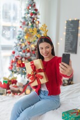 Cute beautiful young asian lady woman wearing reindeer headband selfie with her mobile phone holding gift box posing in front a big Christmas tree with lots of decoration lights gift box and ornaments