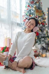 Cute beautiful young asian lady woman wearing earmuffs posing cheerfully in front a big Christmas tree with lots of decoration lights and gift box and ornaments
