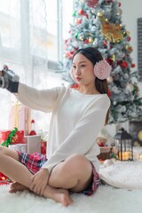 Cute beautiful young asian lady woman wearing earmuffs holding an vintage film camera and posing cheerfully in front a big Christmas tree with lots of decoration lights and gift box and ornaments