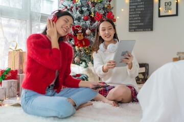 Obraz na płótnie Canvas Two young asian female lady friends in a room listening to new songs on their headphone via their digital tablet celebrate Christmas together 