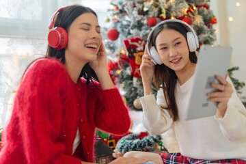 Two young asian female lady friends in a room listening to new songs on their headphone via their digital tablet celebrate Christmas together 