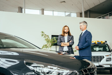Naklejka na ściany i meble Customer in vehicle showroom, male person buying transport, auto dealer business. Business man chosing a new car standing with salesperson in the car showroom.