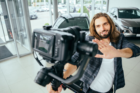 Social media influencer creating content. man shooting video of himself using camera on tripod. Smiling bearded hipster long haired guy communicating with subscribers in automobile showroom.