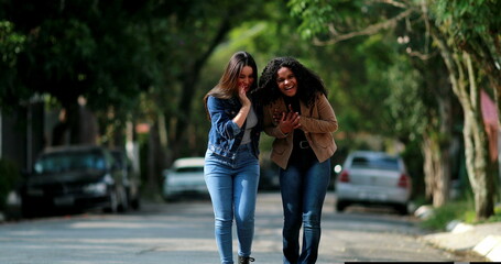 Two beautiful diverse girlfriends walking and laughing together while talking in street, two happy...