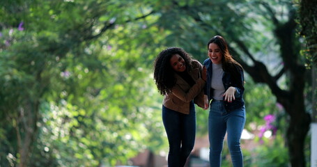Naklejka na ściany i meble Two diverse young women laughing together while walking outside in street