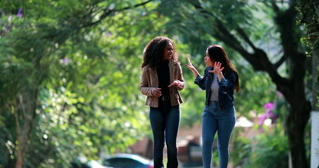 Two diverse young women laughing together while walking outside in street - obrazy, fototapety, plakaty