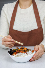 The girl holds a plate of rice noodles and chicken in her hands. Dark home kitchen background