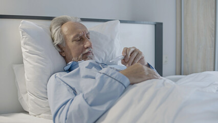 Sick pensioner looking at thermometer, lying in bed, seasonal flu, coronavirus