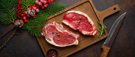 Two raw uncooked meat beef steaks on wooden cutting board with knife and seasonings on dark rustic...