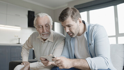 Happy young man and senior father looking through photos on smartphone together, fun