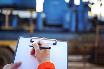 An engineer is writing on paper the list the maintenance detail with a heavy pumping machine as blurred background. Industrial expertise occupation working scene, selective focus at hand's part.	
