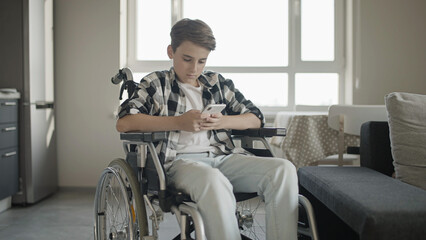 Schoolboy in wheelchair reading messages on smartphone at home, social isolation - obrazy, fototapety, plakaty