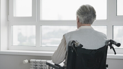 Senior man with disability sitting in wheelchair alone, looking out the window