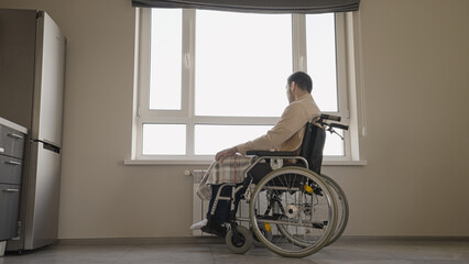 Sad middle-aged man with disability sitting in wheelchair, looking out the window