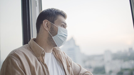 Middle-aged man in protective face mask looking out window, pandemic safety