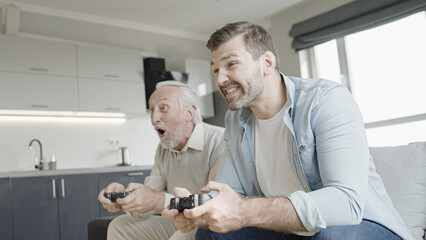 Obraz premium Cheerful senior father and adult son playing video game at home, having fun together