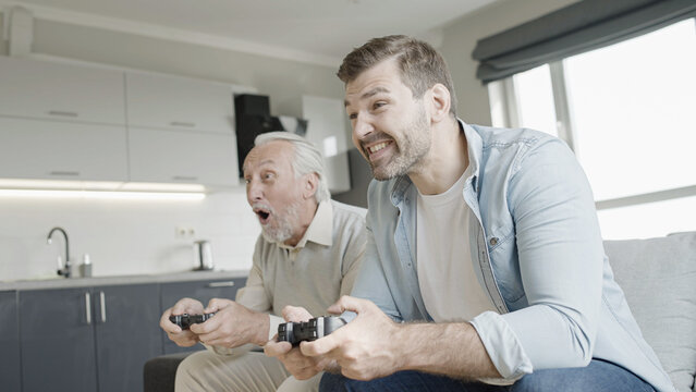 Cheerful senior father and adult son playing video game at home, having fun together