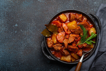 Beef meat stew with potatoes, carrot and delicious gravy in black casserole pot with bay leaves and fresh green herbs with spoon on dark rustic concrete background from above, space for text.