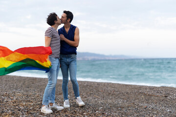  Beautiful gay young couple embraces and holds a rainbow flag. Happy couple enjoy at the beach