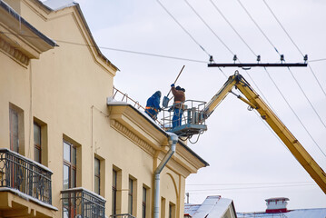 Fototapeta na wymiar Men on roof with shovel clear snow and icicles from roof of an old building. Prevention of snow falling from the roof. City service remove snow from roof. Workers in crane bucket clean snow and ice