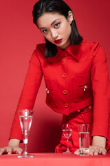 brunette asian woman in elegant jacket standing near glasses with clean water on red background.