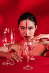 nude asian woman with creative makeup and line on face touching glass with pure water on red background.