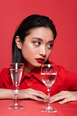 portrait of brunette asian woman with makeup looking away near blurred glasses of water isolated on red.