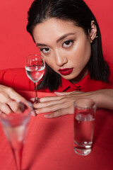 brunette asian woman with makeup and ear cuff looking at camera near glasses with water on blurred foreground isolated on red.