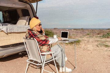 digital nomad woman works on laptop talking by mobile phone while traveling with camper van....