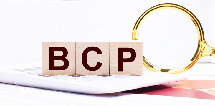Three wooden cubes with the text BCP Business Continuity Plan stand on a white calculator near a magnifying glass. Business concept