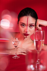 portrait of asian woman with makeup on face divided with line looking at camera near blurred glasses on red background.