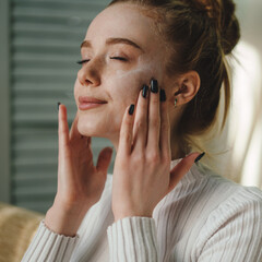Portrait of a young freckled woman with perfect fresh skin applying face cream on her face. Spa and...