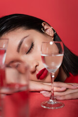 portrait of asian woman with makeup and closed eyes near glasses with pure water on blurred foreground isolated on red.