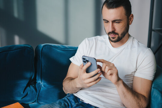 Cheerful Handsome Young Man With Under Eye Patches Using His Smart Phone While Sitting On The Couch At Home. Text Message. Happy Lifestyle.