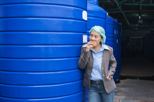 Asian Supervisor Woman Standing Eating Bread With Haste During Lunch Break In Water Tank Warehouse In Processing Factory