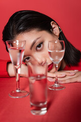 portrait of brunette asian woman with ear cuff and makeup looking at camera near blurred glasses with water isolated on red.